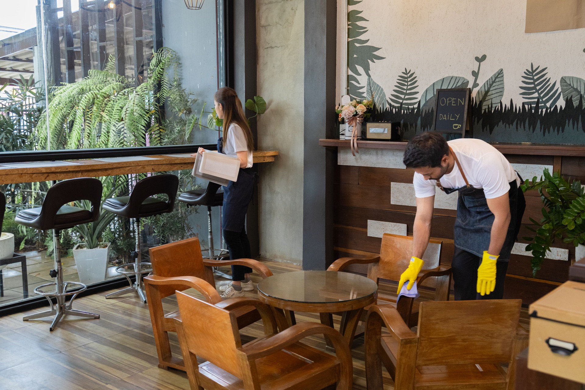 Waiters couple asian or indian wear aprons and cleaning gloves help clean up together, prepare to open shop in morning. Small business owner preparing to support customers come in cafe and restaurant.