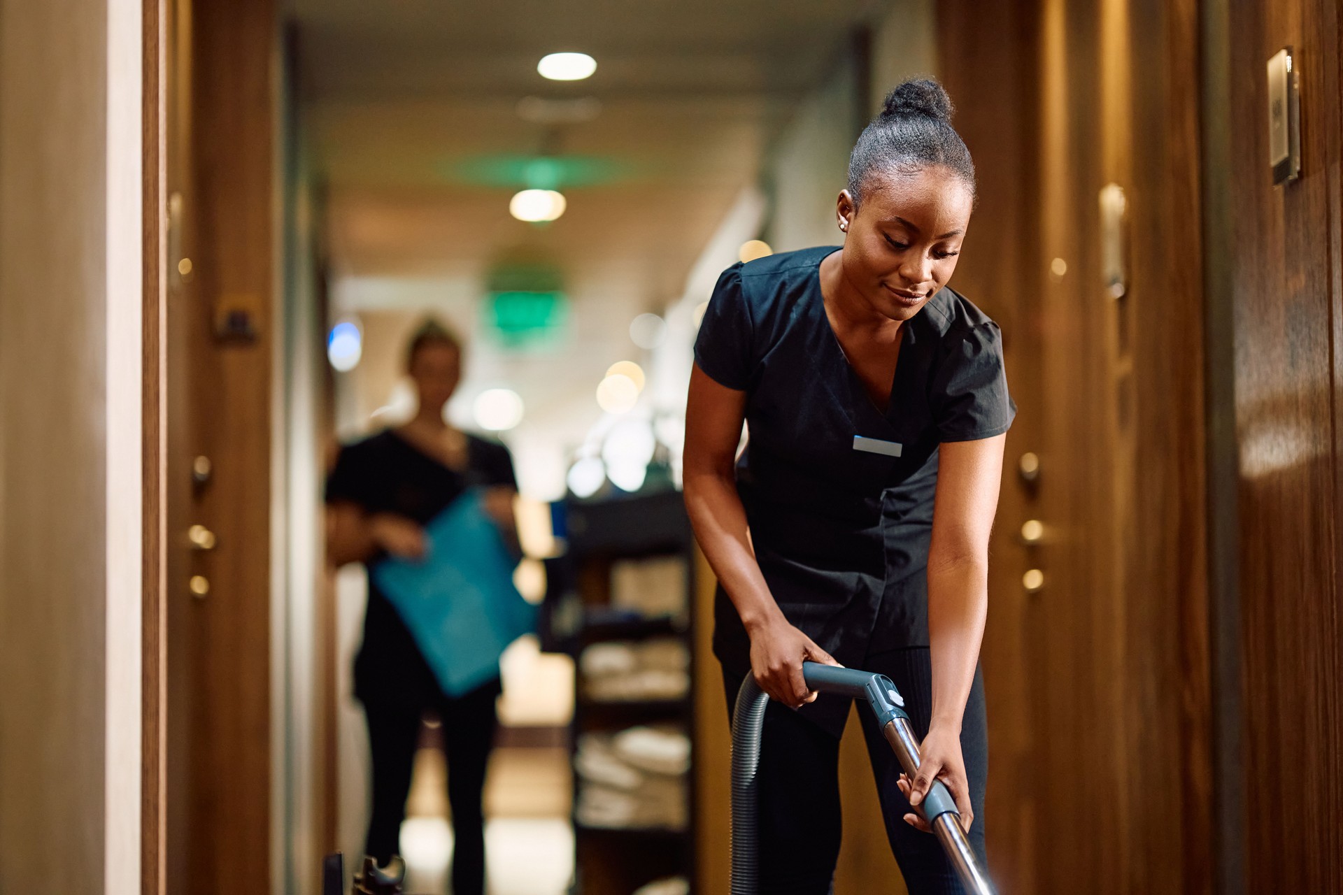 Smiling black maid vacuuming hotel hallway.
